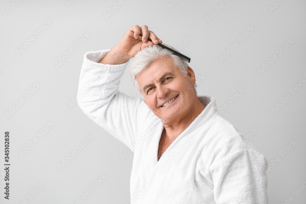 Happy mature man in bathrobe combing his hair on grey background