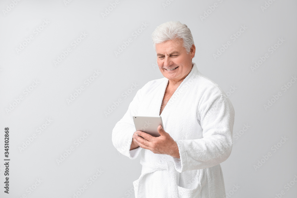 Happy mature man in bathrobe and with tablet computer on grey background