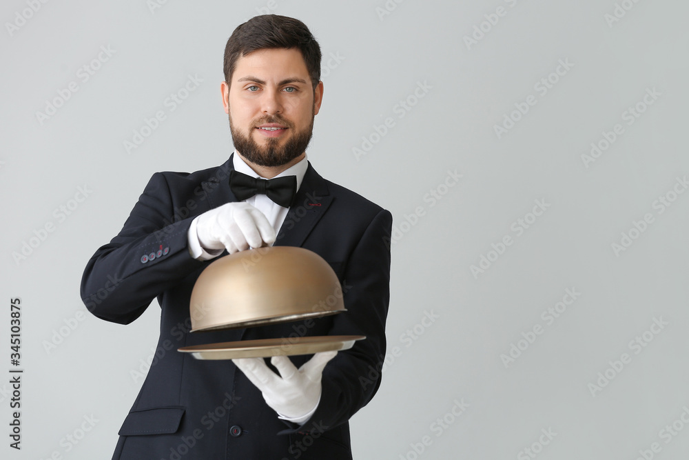 Handsome male waiter with tray and cloche on grey background