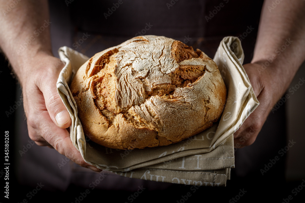 A man is holding fresh rye-wheat bread on a linen towel. Closeup on dark background