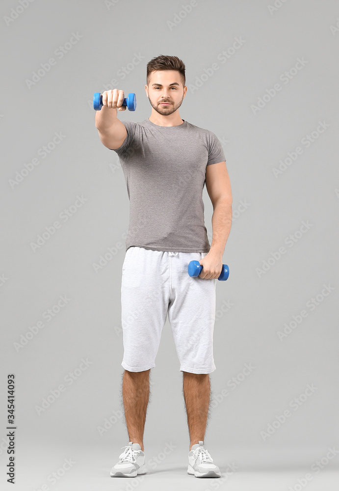 Sporty young man training with dumbbells on grey background