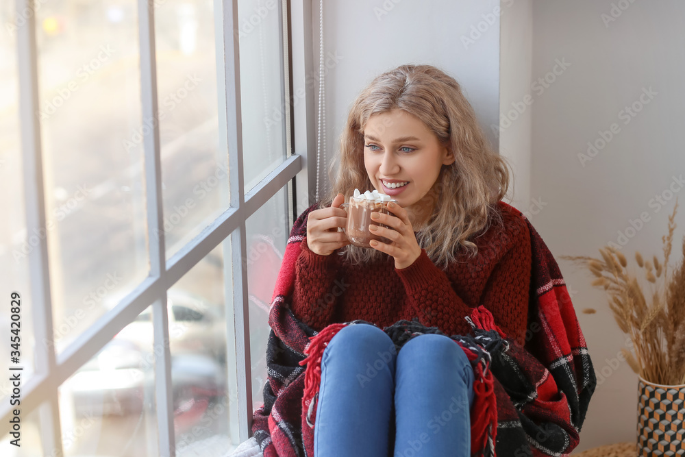 Beautiful young woman drinking hot chocolate near window at home