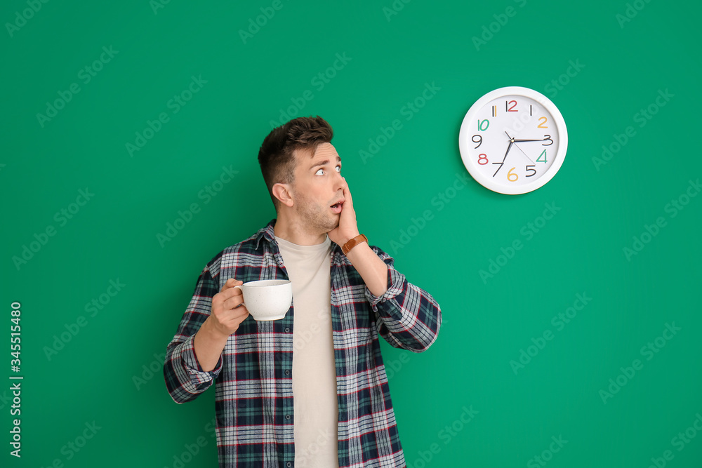 Shocked young man with coffee looking at clock on color background