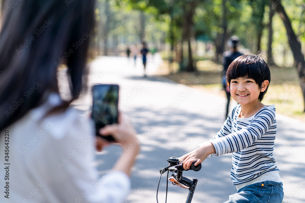 Asian young mother take a photo of kid practicing to ride a bicycle by using smart phone in public p