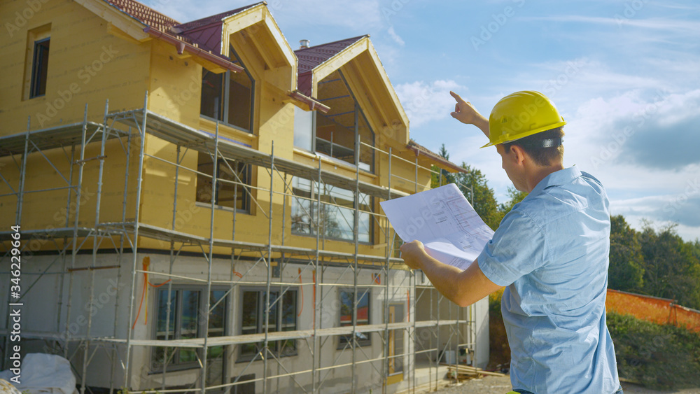 CLOSE UP: Real estate inspector looks at blueprints and points at a house.