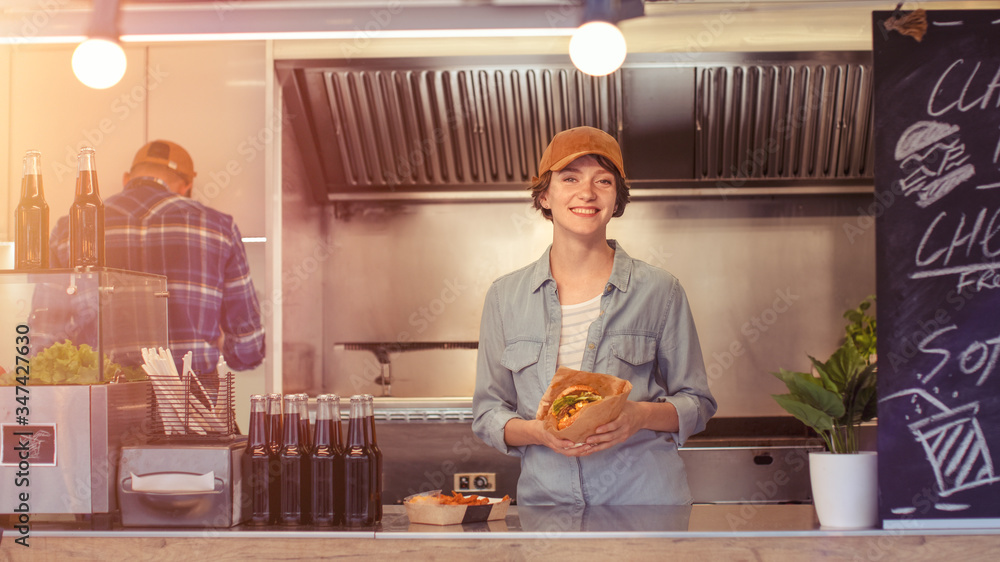 Food Truck Female Employee Hold Tasty Burger, Smiles and Looks at the Camera. Street Food Truck Sell