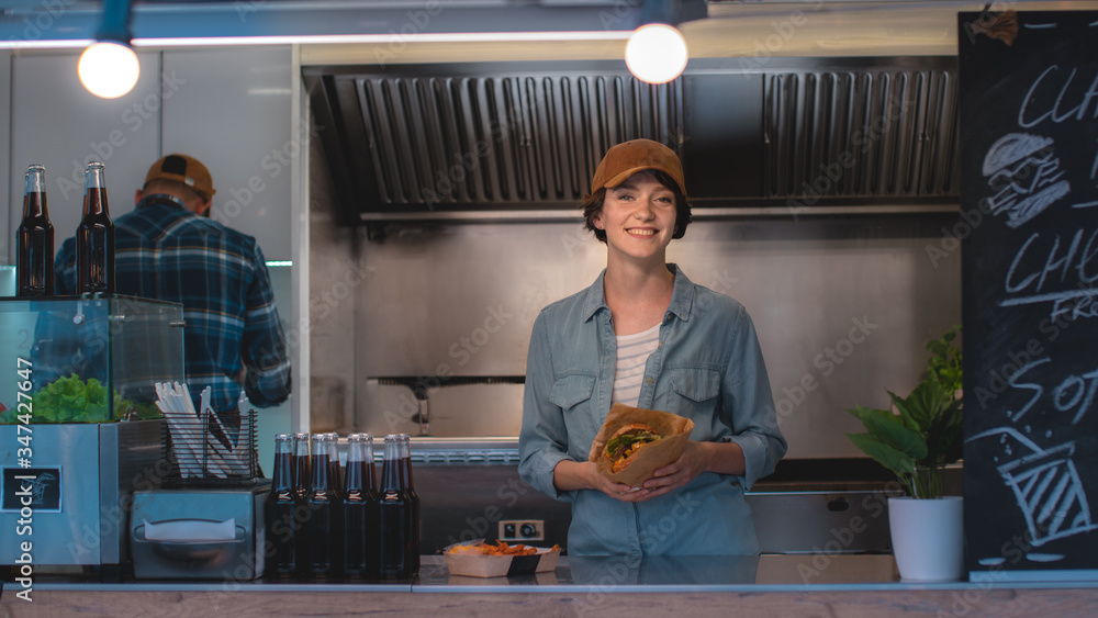 Food Truck Female Employee Hold Tasty Burger, Smiles and Looks at the Camera. Street Food Truck Sell