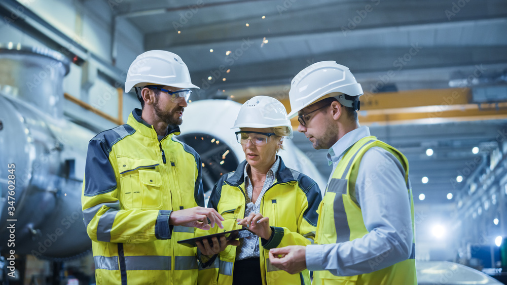 Three Heavy Industry Engineers Stand in Pipe Manufacturing Factory, Use Digital Tablet Computer, Hav