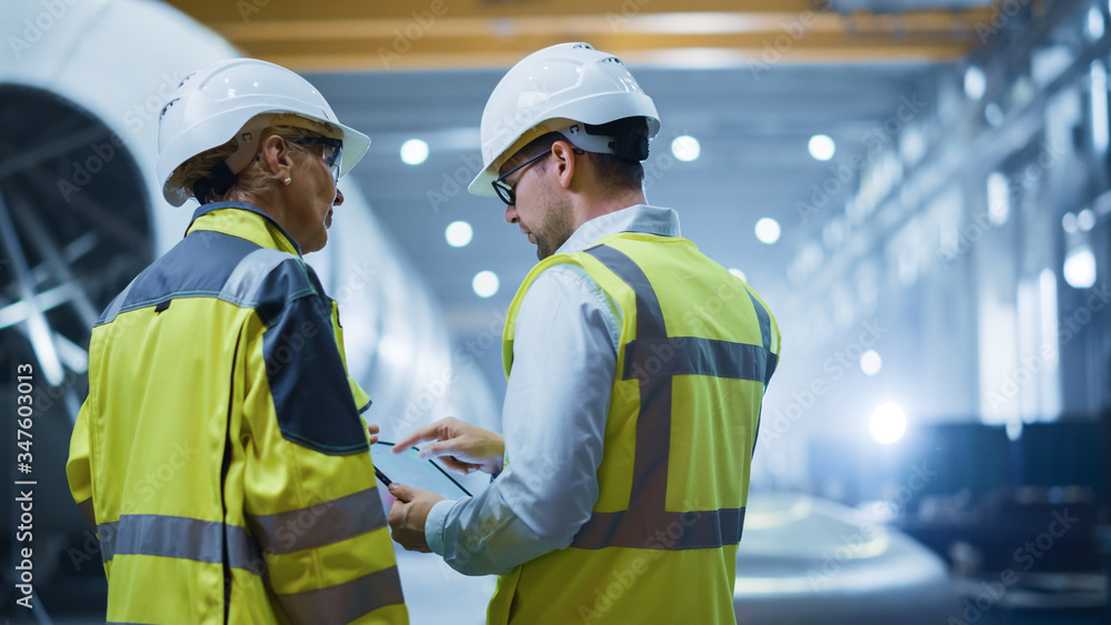 Two Heavy Industry Engineers Stand in Pipe Manufacturing Factory, Use Digital Tablet Computer, Have 