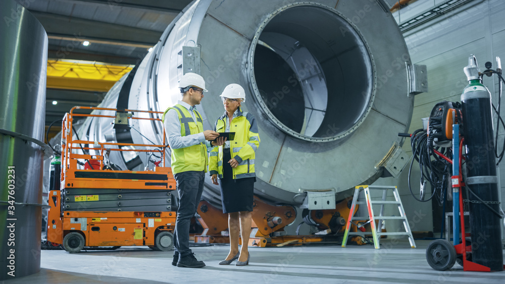 Two Heavy Industry Engineers Stand in Pipe Manufacturing Factory, Use Digital Tablet Computer, Have 