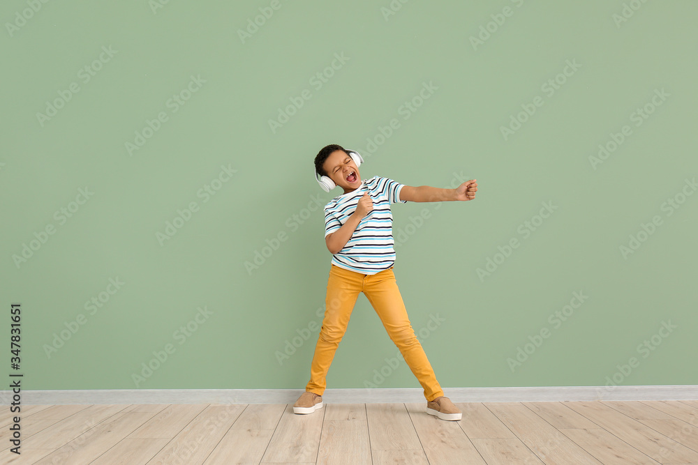 Little African-American boy listening to music and dancing against color wall