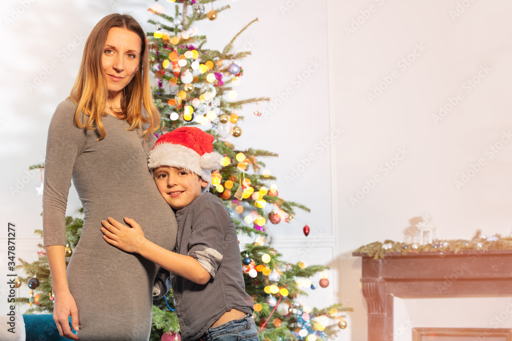 Portrait of mother and little boy wearing Santa hat hug mothers belly near New Year tree