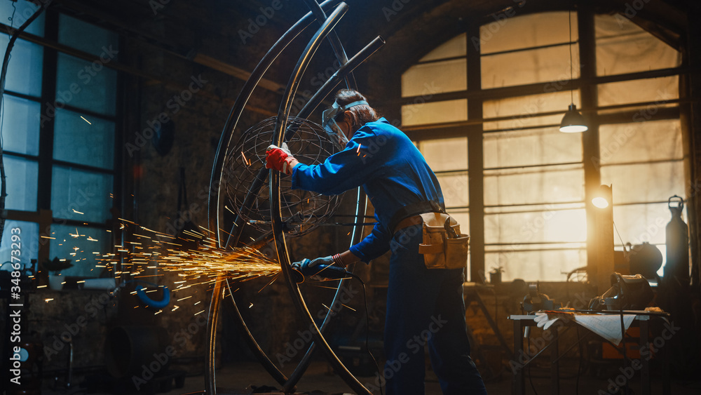 Young Contemporary Female Artist in Blue Jumpsuit is Grinding a Metal Tube Sculpture with an Angle G