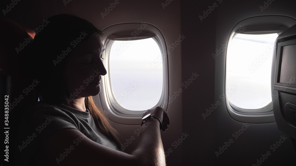 smiling young girl looks at watch to know time against airliner windows in passenger cabin closeup