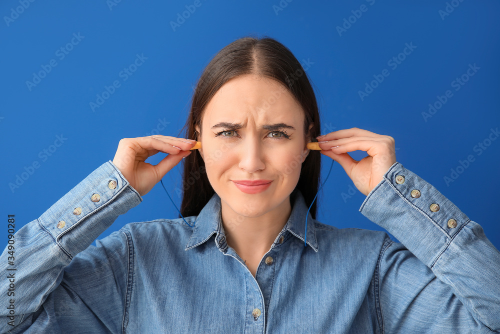 Young woman with ear plugs suffering from loud noise on color background