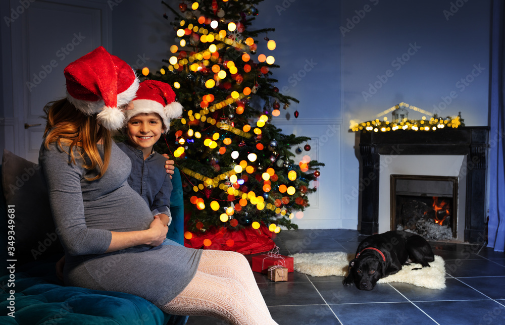 Little boy hold pregnant woman belly sitting on the sofa near New Year tree with dog sleeping by fir