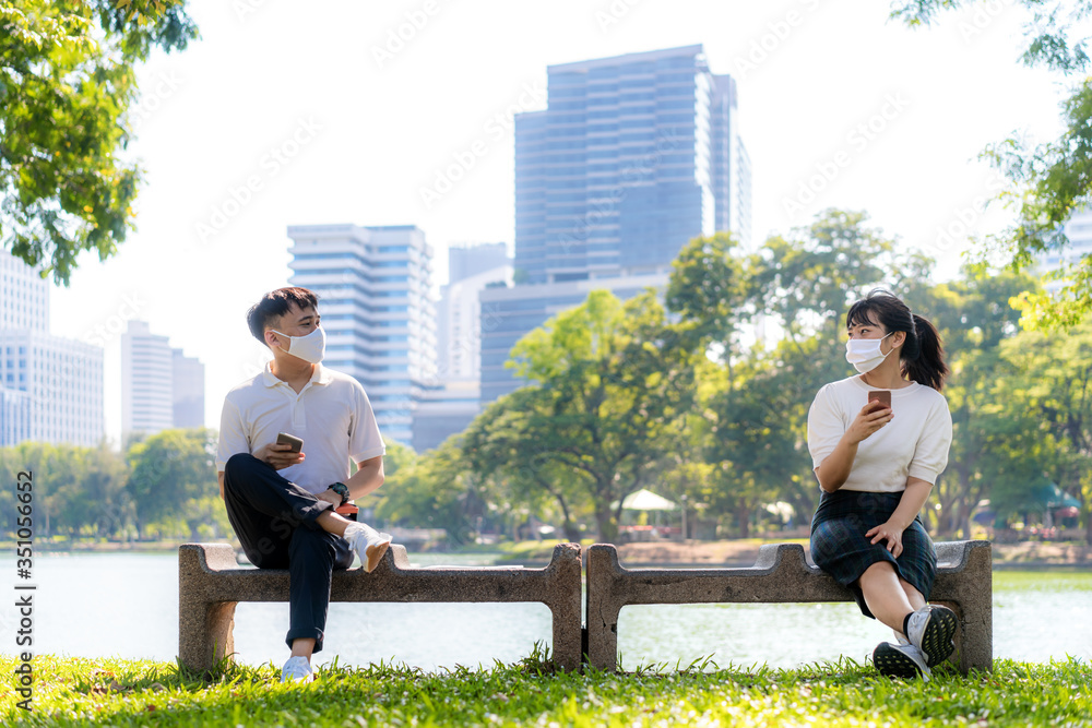 两名亚洲年轻男女在智能手机上聊天，戴着口罩，坐在相距6英尺的地方