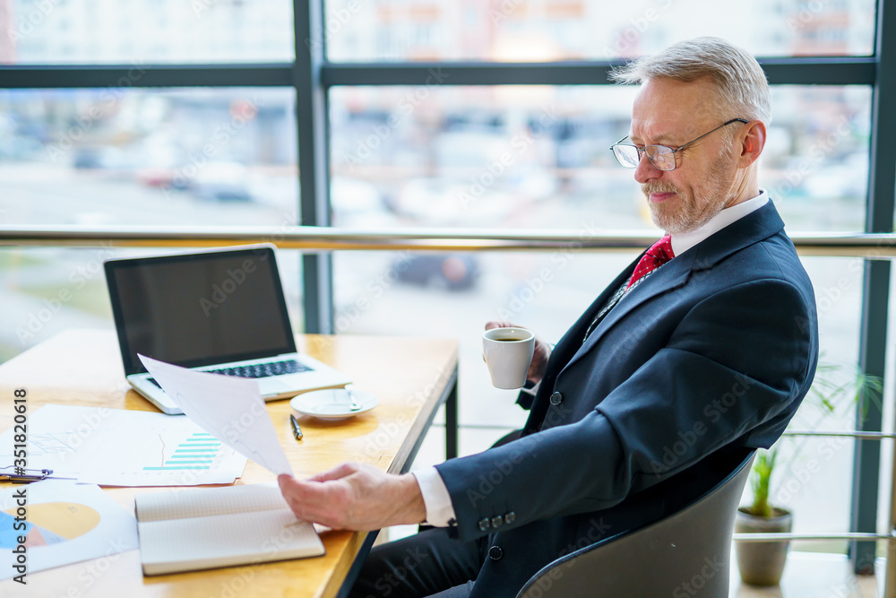 Thoughtful middle aged businessman in suit with a laptop on table while working with documents.