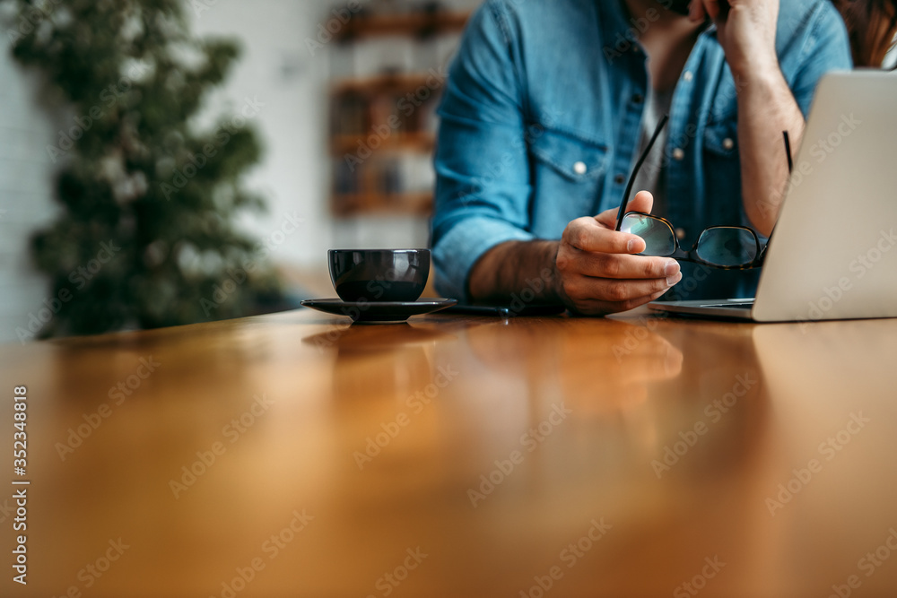 Close-up image of a man sitting at wooden table, copy space. Holding eyeglasses and looking at lapto