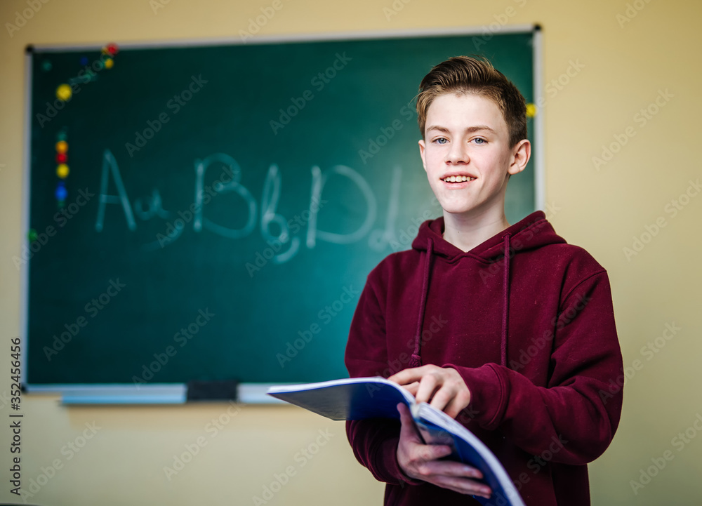 Student of higer school is standing in dark hoodie near the green blackboard in the classroom readin