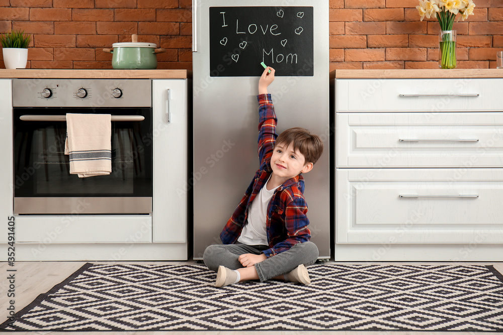 Little boy writing text I LOVE MOM on chalkboard in kitchen