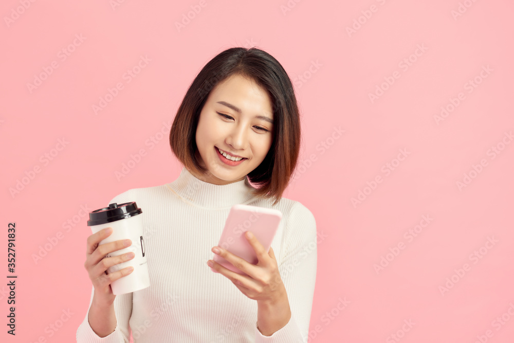 Attractive young Asian woman using mobile phone while holding coffee cup over pink background.