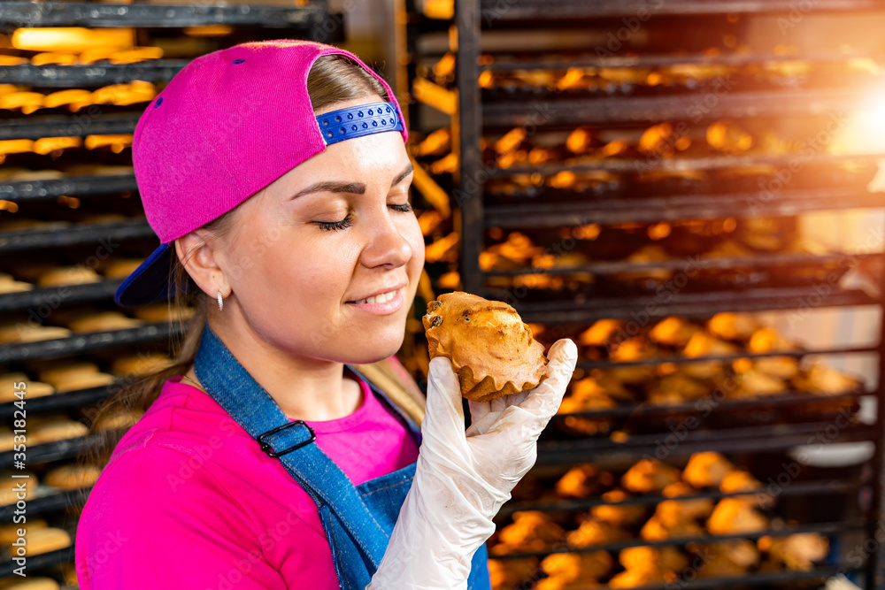 Young happy female worker in sterile cloths holding freshly baked muffins on tinplate inside food pr