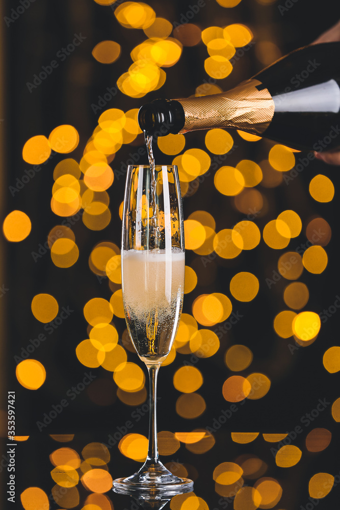 Pouring of champagne in glass on dark background with defocused lights