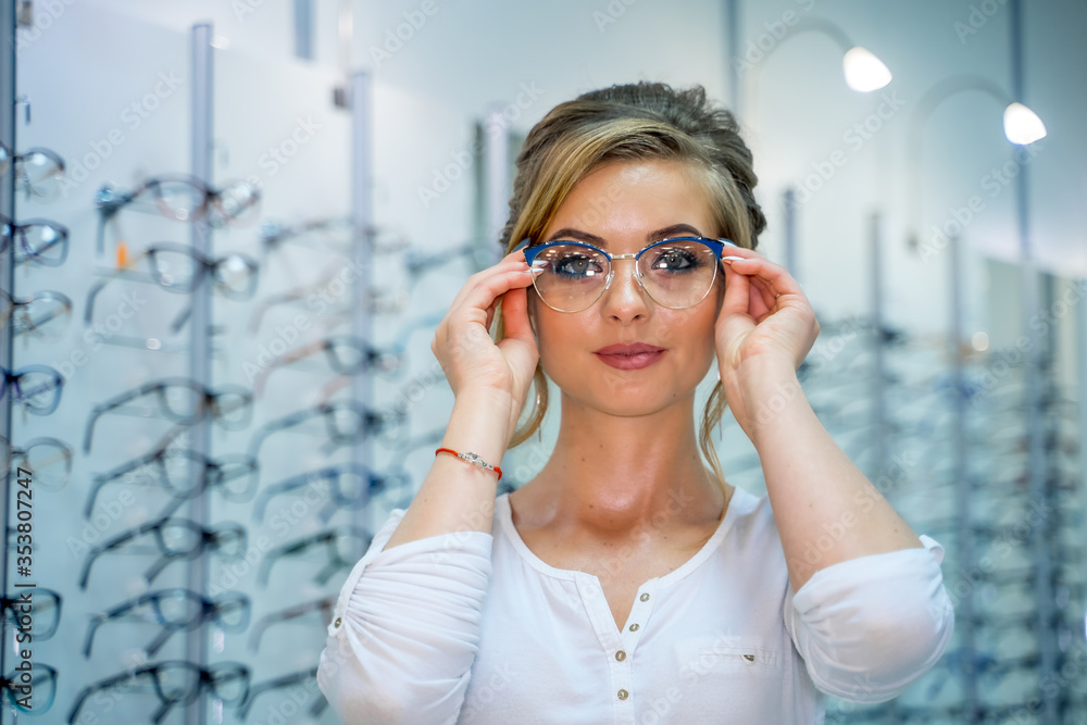 Female is standing with raw of glasses in background in optical shop. Stand with spectacles. Eyesigh