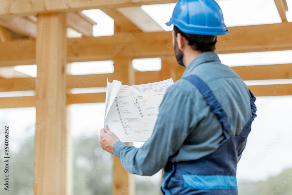 Builder in blue overalls and hard hat with blueprints on the construction site. Building wooden fram