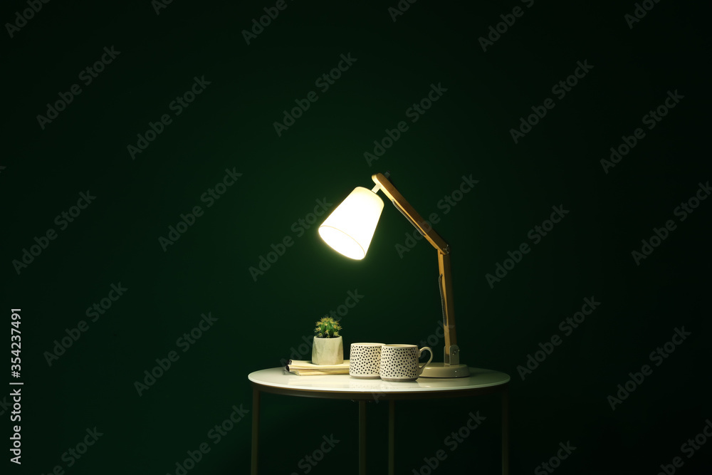 Glowing lamp, cups and houseplant on table in dark room