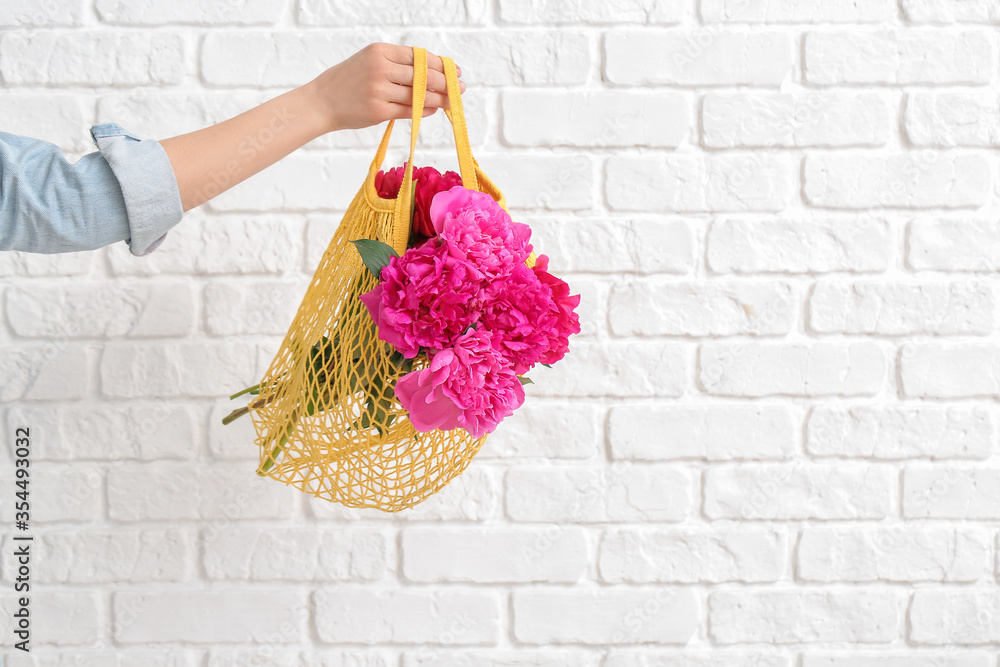 Female hand with beautiful peony flowers in bag on white brick background