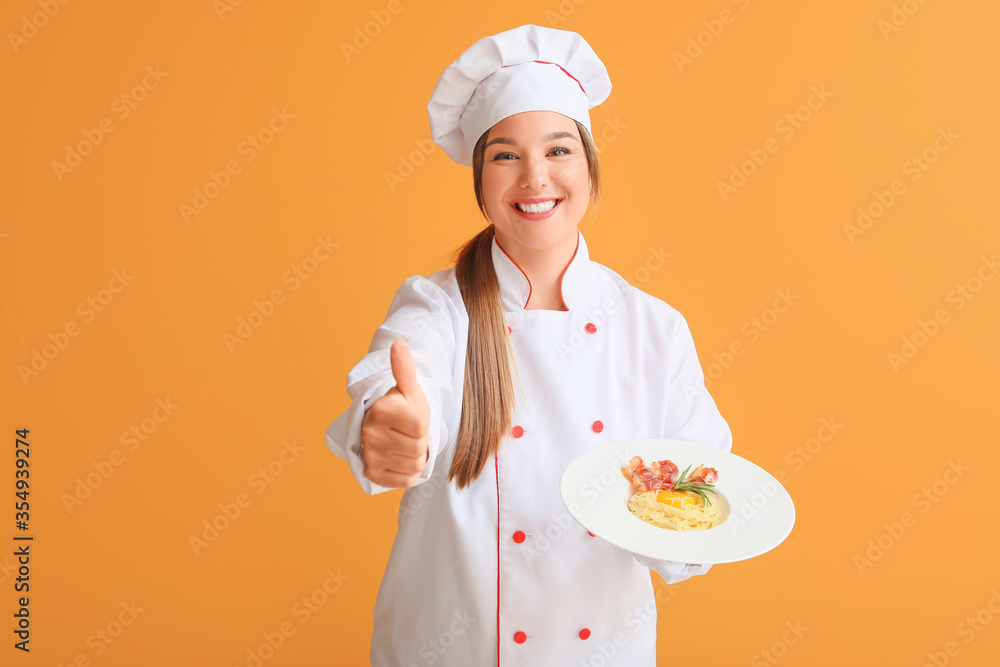 Young female chef with tasty dish showing thumb-up on color background