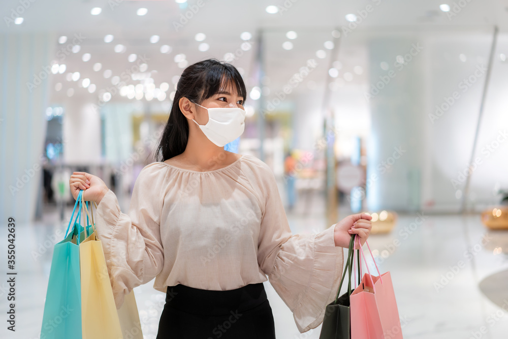 一名亚洲女子戴着口罩在购物中心散步，手里拿着健康购物袋