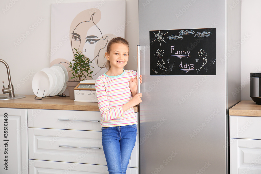 Little girl near chalkboard on refrigerator in kitchen