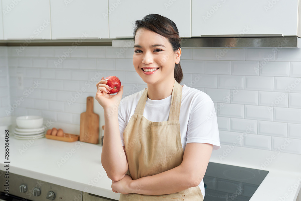 Asian women wear apron posing in the kitchen at home looking at camera, red apple in hand