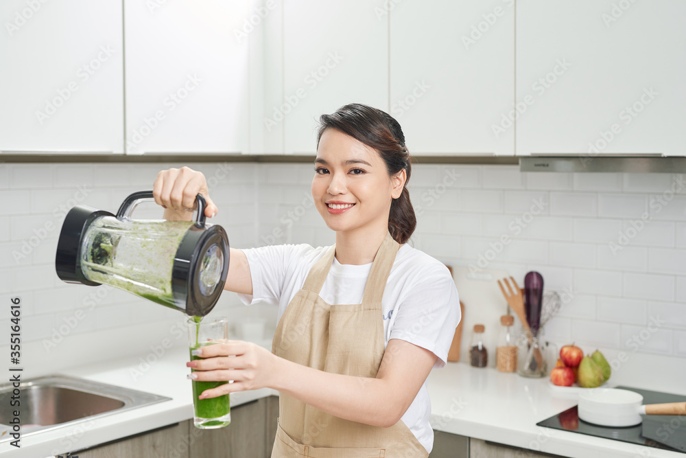 穿着亚麻围裙的年轻女性将绿色奶昔饮料从搅拌机倒到装满蔬菜的瓶子里