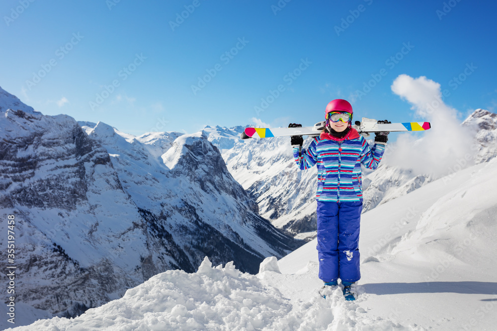 年轻的微笑女孩穿着五颜六色的衣服，站在雪地里，戴着粉色头盔和彩色眼镜，滑雪