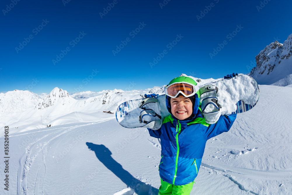 可爱的微笑小男孩把滑雪板扛在肩上，摘下口罩，露出灿烂的笑容。