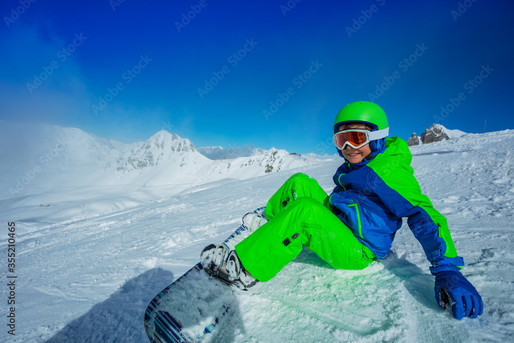 Happy boy on snowboard sit on the snow slope in full sport equipment helmet and mask