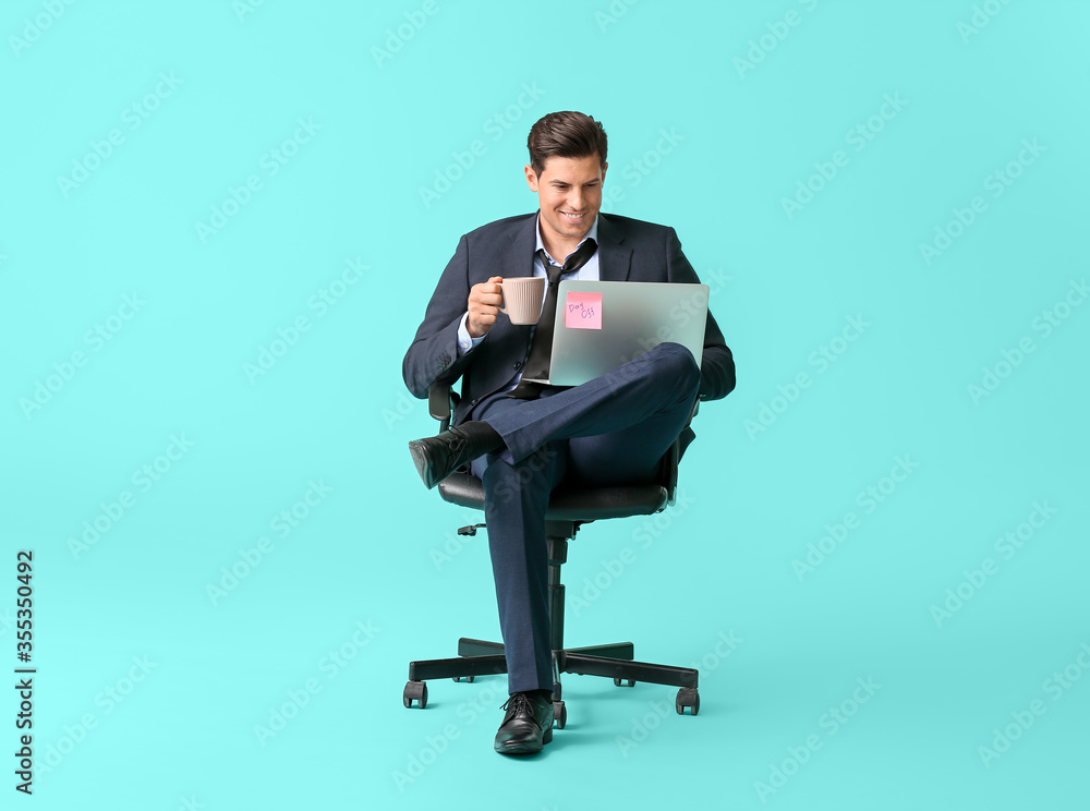 Young businessman with laptop and coffee relaxing on chair against color background