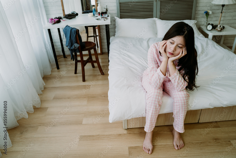 gloomy chinese lady sitting at the edge of bed is having monday blue and doesn’t want to go to work.