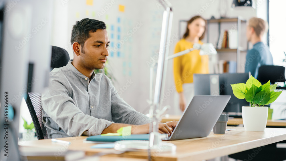 Smart and Handsome Indian Office Worker Sitting at His Desk works on a Laptop. In the Background Mod