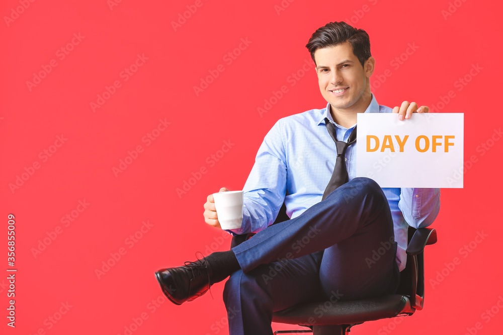 Young businessman holding paper with text DAY OFF and drinking coffee against color background