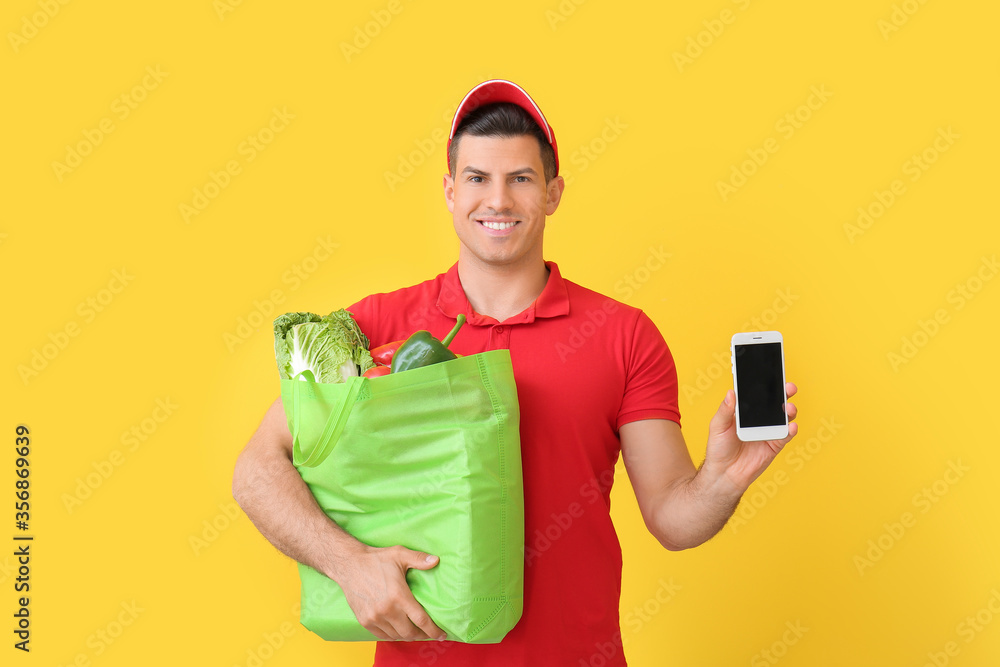 Delivery man with food in bag and mobile phone on color background