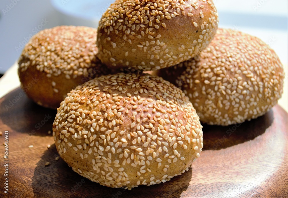 Ruddy rye homemade buns with sesame seeds on a flat wooden plate, bathed in beams of bright sun (on 