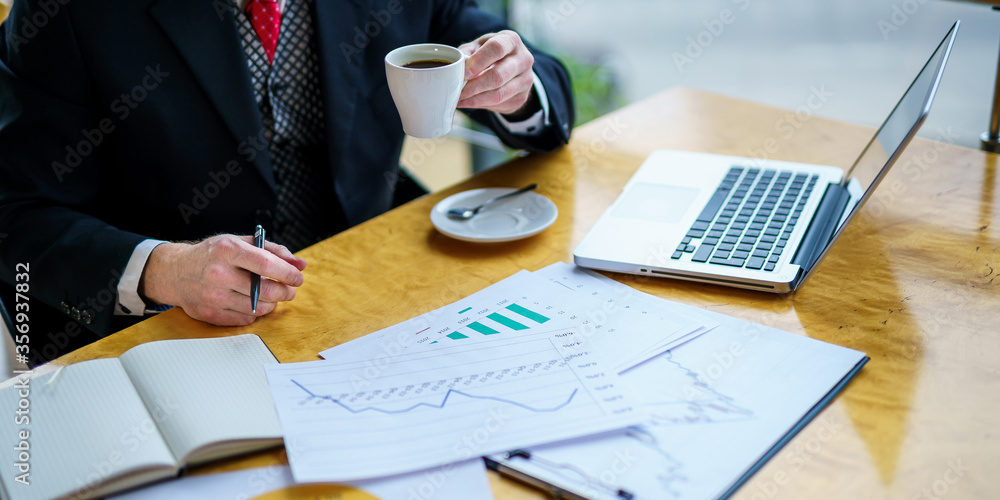 Senior businessman wears white shirt and dark suit. Drinking coffee at table with documents on it. B