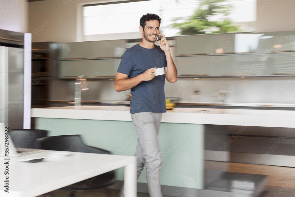 Smiling man drinking coffee and talking on cell phone in kitchen