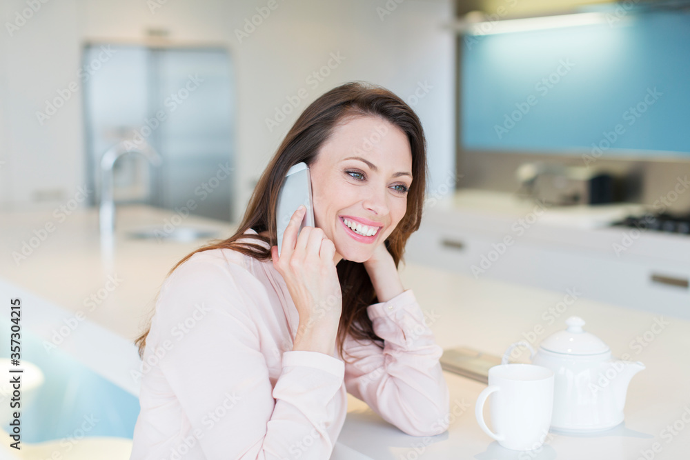 Smiling woman talking on phone in kitchen, tea cup tea pot on counter