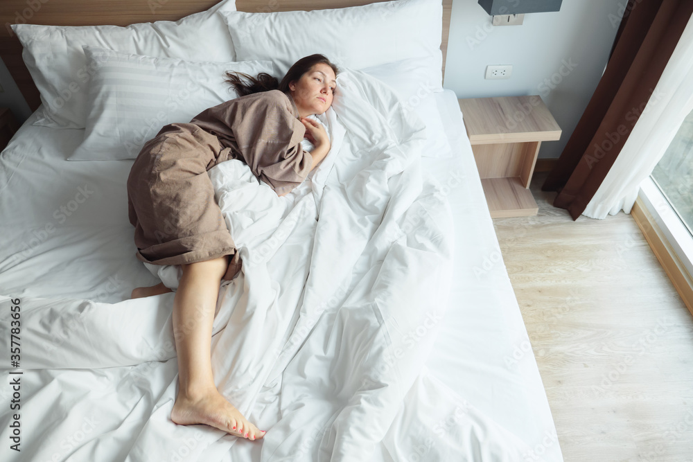 cheerful woman in beige bathrobe rests lying on soft bed with blanket and pillows and yawns stretchi